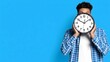 © lucas - Man holding a clock in front of his face with a blue background, time management and deadline concept