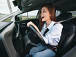 © SHOTPRIME STUDIO - A woman is in the driver's seat of a car with her hands on the steering wheel, mouth wide open, appearing shocked or surprised