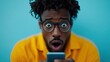 © Maximages  - A young man in glasses and a yellow shirt looks shocked as he views his phone. His wide-eyed expression and the blue background emphasize the magnitude of his surprise.