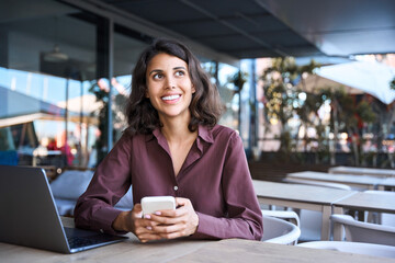 Canvas Print - European business woman manager using phone mobile app sitting outdoors at cafe office. Latin Hispanic young female businesswoman freelance working on smartphone, looking aside dreaming. Copy space