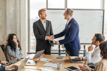 Aufkleber - Two businessmen in suits are shaking hands in a modern office, while other colleagues look on. Large screen behind them displaying a graph, and laptops and other work materials are on the table.