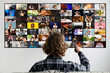 © Prostock-studio - This image shows a man sitting in front of a large television screen. The television is displaying a grid of multiple small videos. The man is holding a remote control in his right hand