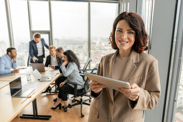 Aufkleber - A businesswoman in a beige blazer stands in a modern office, holding a tablet and smiling at the camera. She is positioned near a large window with a view of a cityscape.