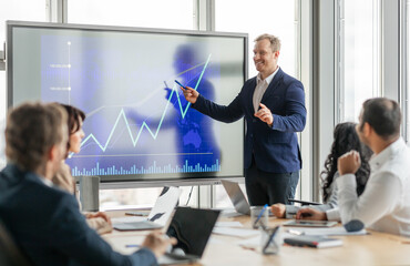 Poster - A businessman stands at the front of a meeting room, pointing at a large screen displaying a financial graph. He is presenting data to his colleagues, who are seated around a conference table.