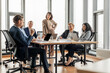 © Prostock-studio - A group of business professionals are having a meeting in a modern office. The meeting is taking place around a large, rectangular table with several laptops and papers on it.