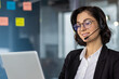 © Liubomir - Smiling businesswoman wearing headset working on laptop in office setting. Professional appearance, engaging in customer support conversation. Office environment with sticky notes on glass board