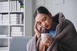 © amnaj - Young asian woman feeling sick having headache holding a mug with hot drink sitting on a sofa at home