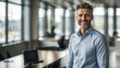 © Najam - A close-up portrait of a happy businessman standing in an office with large windows, natural light pouring in, his expression bright and confident.