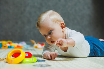 A little six-month-old boy is learning to lie on his stomach. The baby plays with toys lying on the floor.