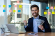 © Liubomir - Businessman celebrating financial success holding cash at desk with laptop and office supplies.