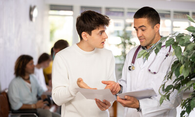 Wall Mural - Young male doctor and patient discussing medical document standing in waiting room