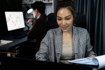 Businesswoman with stressful face looking document paperwork file while coworker trading stock market at back at over late night time at modern office, working on startup company project. Infobahn.