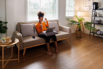 Image of cheerful excited happy african american woman afro hair,  using laptop while sitting on chair in living room