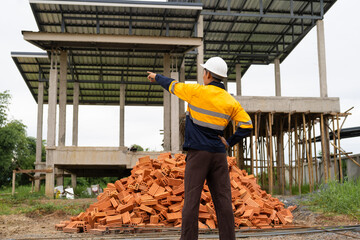 Wall Mural - A male architect wearing a hard hat and safety vest checks his laptop while holding a clipboard with house plan papers. He reviews blueprints, construction documents,house structure details on-site.