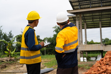 Two male architects wearing hard hats and safety vests discuss construction design on-site. collaborating with the contractor on the house structure, including iron framework, brickwork, and roofing.