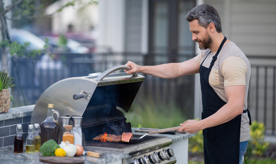 Poster - Hispanic man cooking on barbecue in the backyard. Chef preparing barbecue. Barbecue chef master. Man in apron preparing delicious grilled barbecue food, bbq meat. Grill and barbeque.