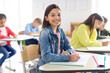 © Home-stock - Happy European schoolgirl sitting at desk in classroom and smiling at camera, writing in notebook. Group of classmates studying on background