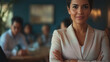 © VikaKa - A cheerful businesswoman standing confidently with her arms folded. The background shows her team sitting at a table, actively discussing