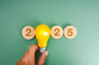 © tete_escape - Happy new year 2025 innovation technology, creative market trend, inspiration and idea concept. 2025 calendar number on wood blocks with glow yellow light bulb holding in man hand on green background.
