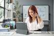 © amnaj - Young asian businesswoman smiling and talking on the phone while working on a digital tablet in the office