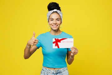 Wall Mural - Young shocked Latin woman wears blue top t-shirt headband casual clothes hold gift certificate coupon voucher card for store show thumb up isolated on plain yellow background studio Lifestyle concept