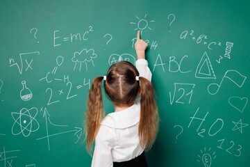 Back rear view little fun school girl 7-8 years old pony tails wear white shirt uniform point index finger on symbol isolated on green blackboard background Childhood kids education lifestyle concept