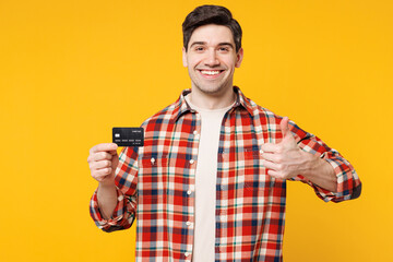 Sticker - Young smiling happy man wears red checkered shirt casual clothes hold in hand mock up of credit bank card show thumb up isolated on plain yellow orange background studio portrait. Lifestyle concept.