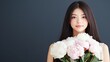 © lililia - A woman with long hair holds a bouquet of pastel-colored peonies and roses, looking warmly at the camera in a contemporary studio with a dark gray backdrop