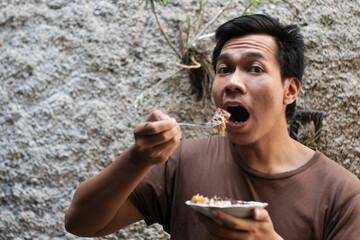 Cheerful A Southeast Asian man holding fork and white plate, with tasty grilled banana looking smiling at camera on grey wall. Portrait of happy man.