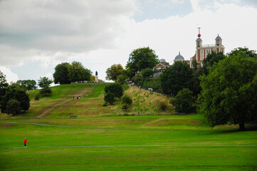 A scenic view of a green hill with walking paths and people, set against a backdrop of historical architecture under a partly cloudy sky.