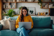 © Instacraft.Studio - A young woman is sitting on a couch in her living room, working on her laptop. She is smiling and appears to be focused on her work.