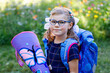 © Irina Schmidt - little girl with glasses and a blue backpack, dressed in a navy uniform, holds a colorful school cone called Schultuete and backpack. Happy child smiling, ready for her first day of school