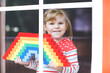 © Irina Schmidt - A smiling girl holds a rainbow of colorful building blocks behind a window. She wears a red polka-dot shirt over a striped one, with her blonde hair tied back.