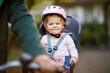 © Irina Schmidt - A smiling toddler girl wearing a pink helmet sits securely in a bicycle child seat, ready for a ride. The background is blurred, focusing on the child joyful expression
