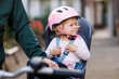 © Irina Schmidt - A smiling toddler girl wearing a pink helmet sits securely in a bicycle child seat, ready for a ride. The background is blurred, focusing on the child joyful expression