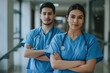 © Alberto Gonzalez  - Two confident healthcare professionals, a man and a woman, wearing blue scrubs and stethoscopes, standing with crossed arms in a hospital hallway.