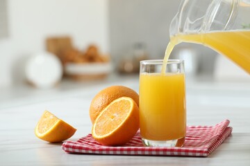 Wall Mural - Pouring tasty orange juice from jug into glass at white table with citrus fruits