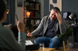 © Mediaphotos - Hispanic man sitting in office talking to professional therapist while holding head in hand, indicating stress and frustration. Environment includes books, desk, and comfortable seating