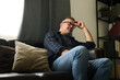 © Mediaphotos - Man relaxing on couch in living room, resting head on hand while considering thoughts with serious expression near window with natural light coming through curtain