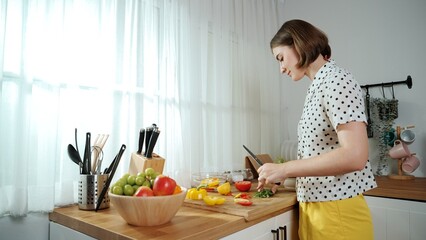 Wall Mural - Energetic mother making salad for breakfast at modern kitchen while chopping vegetable surrounded with kitchenware. Smart female house keeper cooking and preparing food. Healthy lifestyle. Pedagogy.