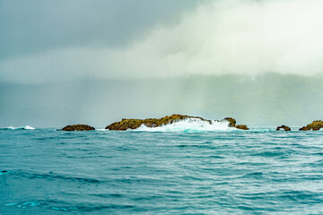 Waves crashing against a rocky outcrop in the ocean with a misty, forested mountain in the background under a cloudy sky. High-quality photo. Uvita Puntarenas Province Costa Rica