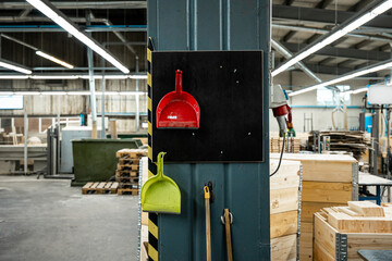 A red and green dustpan hanging on a wall in an industrial factory setting, with wooden pallets and machinery in the background