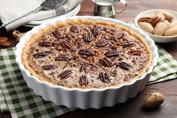 Wall Mural - Delicious pecan pie in baking dish and fresh nuts on wooden table, closeup