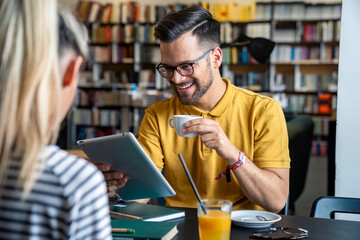 A joyful Caucasian male student holding coffee chats with a female counterpart over a tablet amongst stacks of books, both enjoying beverages in a studious library setting.