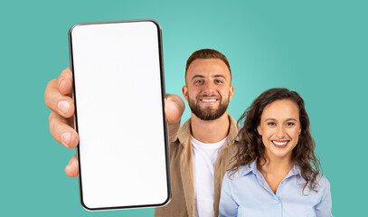 Fototapete - A man and a woman are standing together against a teal background. The man is holding a smartphone with a blank screen towards the camera. They are both smiling and looking directly at the camera.