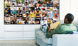 © Prostock-studio - African American man is sitting on a white couch in a living room, watching television. The TV screen is filled with a diverse mosaic of smaller images, showcasing various activities and people