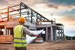 © digitalsync - Construction Project Blueprint: A construction worker in a safety vest and hard hat stands on a construction site, reviewing blueprints against the backdrop of a partially built house under a dramatic