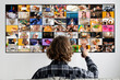 © Prostock-studio - A person sits on a couch facing a television screen, holding a remote in their hand. The television is displaying a grid of colorful images, each showing a different scene, person, or object