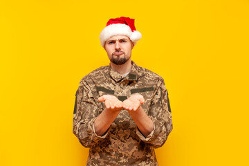 Sticker - young ukrainian army soldier in camouflage pixel uniform and santa claus hat begging and holding empty hands on yellow isolated background, sad ukrainian military cadet asking and giving for new year