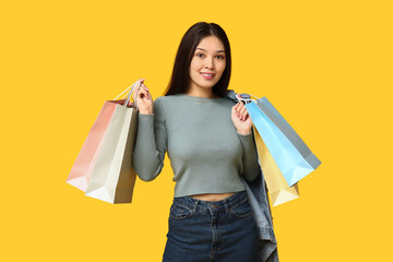 Fashionable young Asian woman in stylish denim clothes with shopping bags on yellow background